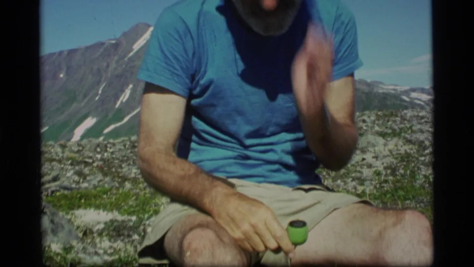 Adult man in a blue T-shirt and khaki shorts sits on an Alaska mountain slope in supplied 1977 home-movie footage.