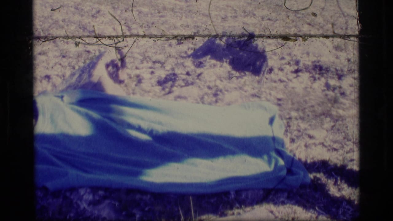 Man lies on a blue blanket on rocky ground in strong sunlight.