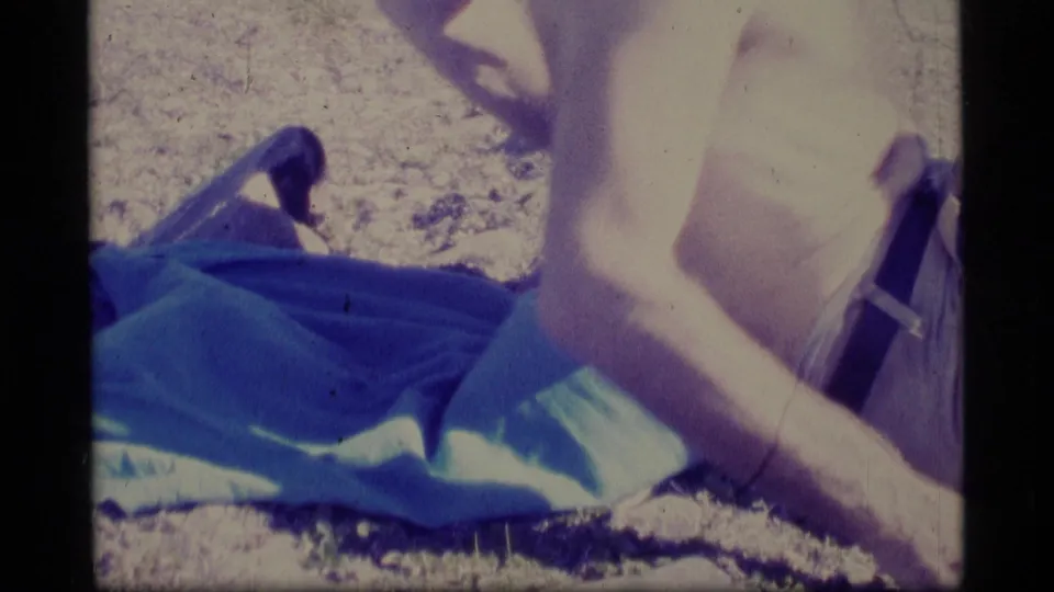 A shirtless adult man reclines on a blue blanket in the Alaska mountain setting, with bright 8mm exposure and color shift.