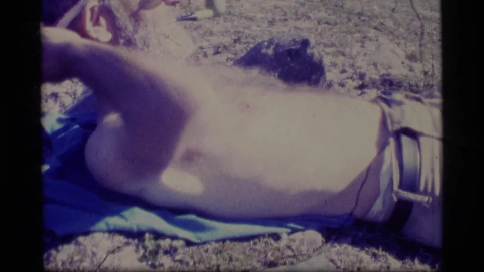 The man lies sideways on a blue blanket in the supplied 1977 Alaska home-movie footage, framed by rocky ground and film borders.