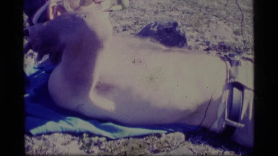 Adult man in a cap sunbathing on a blue towel beside an Alaskan waterway in 1977 home-movie footage.