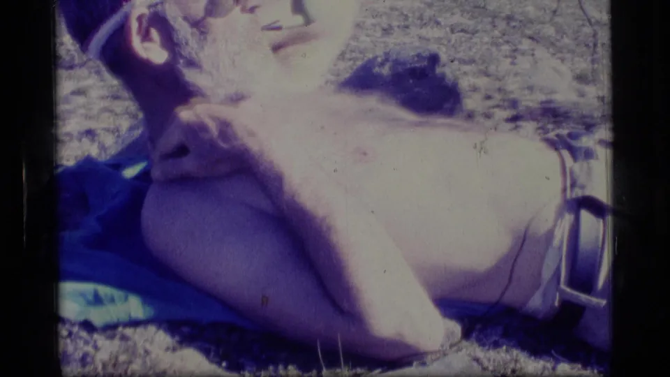 The reclining man rubs or holds his neck while resting on a blue towel in 1977 Alaska home-movie footage.