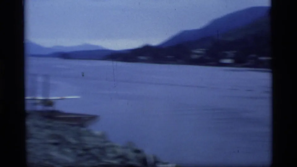 A handheld view across an Alaskan waterway shows mountains, shoreline, and a small boat passing near the foreground.