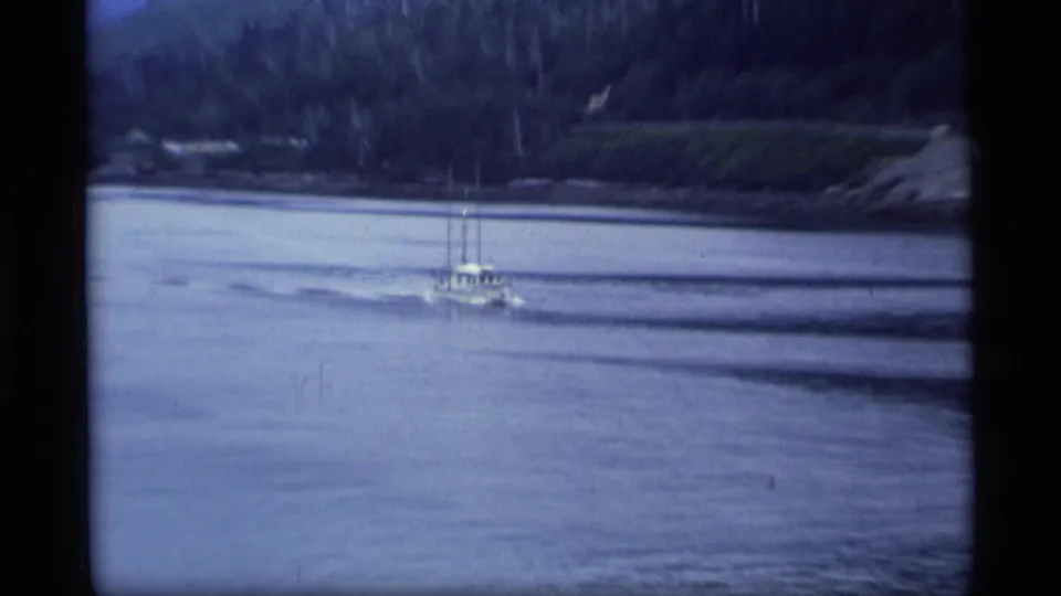 A small boat travels across an Alaskan waterway below a dark wooded shore in 1977 home-movie footage.