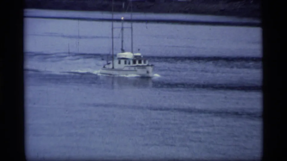 1977 Alaska home-movie frame showing a small motorboat crossing a broad river with wake trailing behind it.
