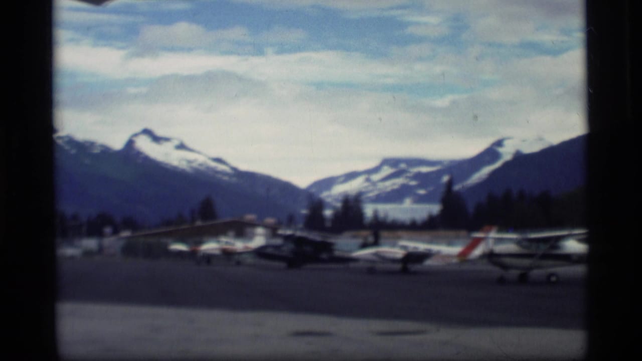 Small airplanes parked on a tarmac with snow-capped mountains behind them.