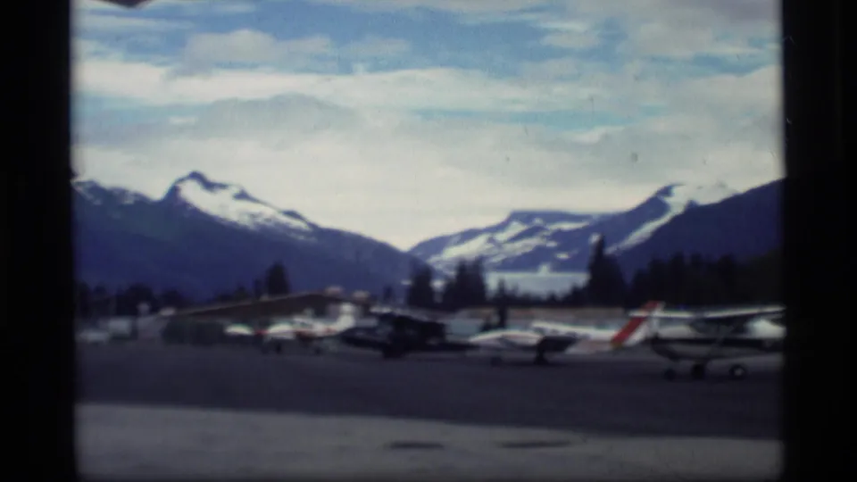 1977 Alaska airfield view with parked small aircraft on tarmac beneath snow-capped mountain peaks.