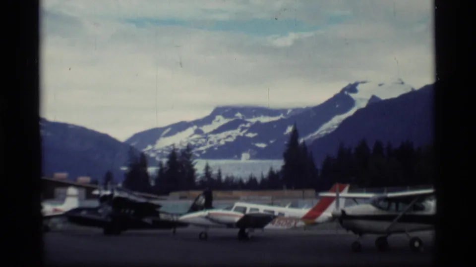1977 Alaska home-movie view of small aircraft parked near a treeline, with snow-covered mountains in the background.