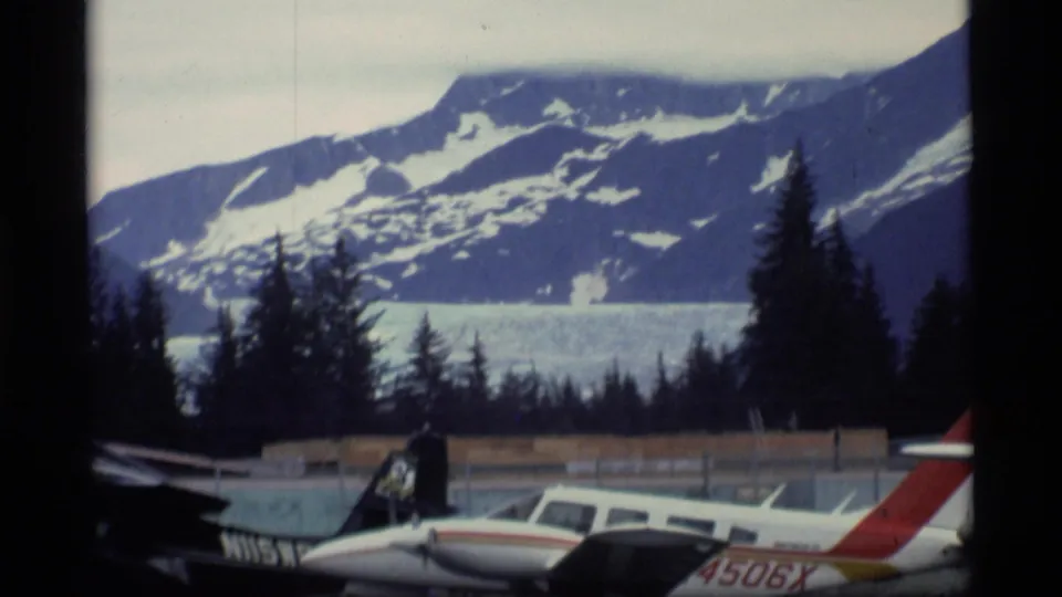 1977 Alaska airfield frame showing parked light aircraft, visible tail markings, dark trees, and snowy mountain terrain.