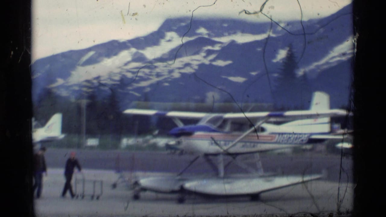 1977 Alaska home-movie frame showing a small floatplane on an airfield, with mountains, trees, film scratches, and a person nearby.