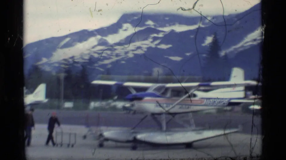 1977 Alaska home-movie frame showing a small floatplane on an airfield, with mountains, trees, film scratches, and a person nearby.