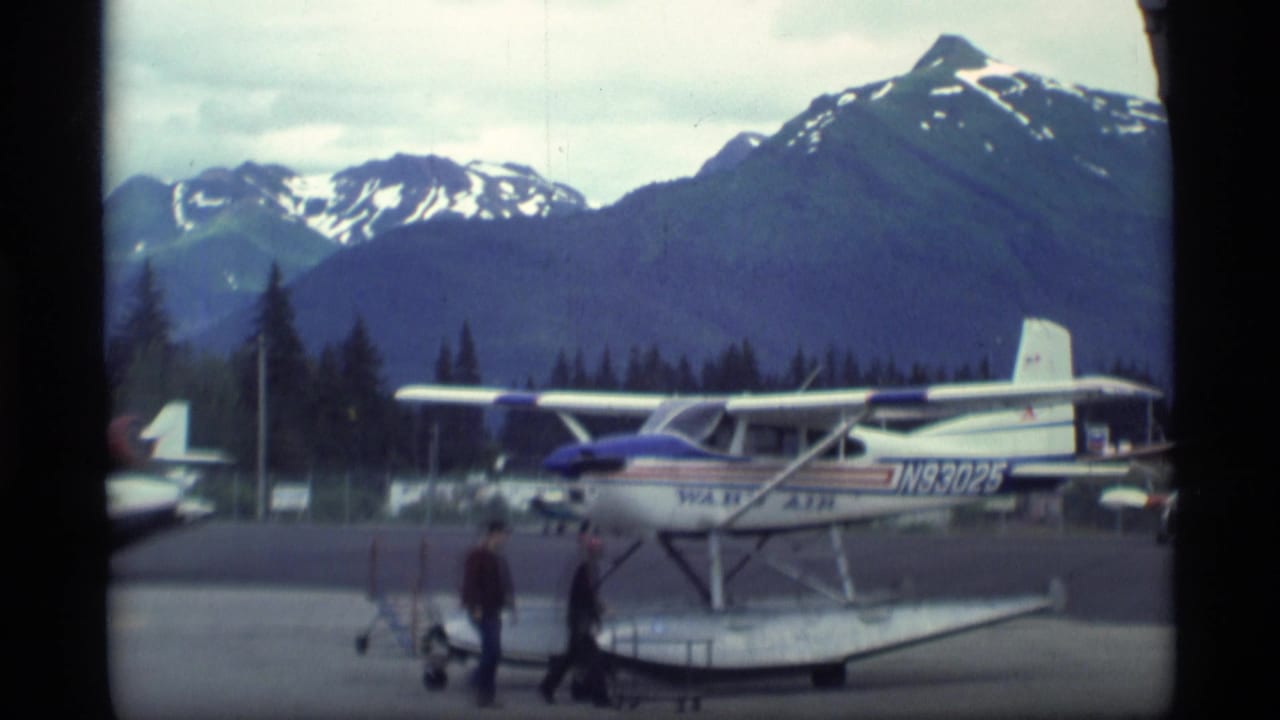 1977 Alaska frame with two adults walking near a marked floatplane, backed by forested slopes and snow-capped mountain peaks.