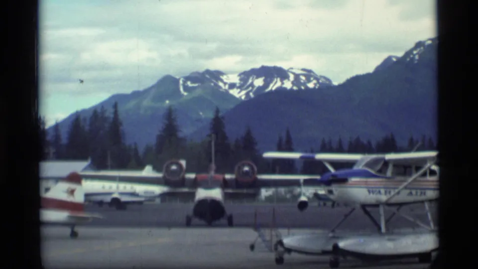 1977 Alaska airfield view with multiple light aircraft, including a floatplane, parked below evergreen trees and snow-patched mountains.