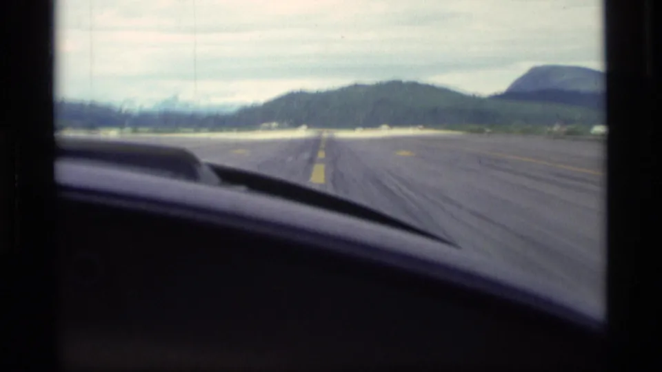 1977 Alaska cockpit-view frame looking down a runway toward low wooded hills, with the aircraft dashboard and windscreen visible.