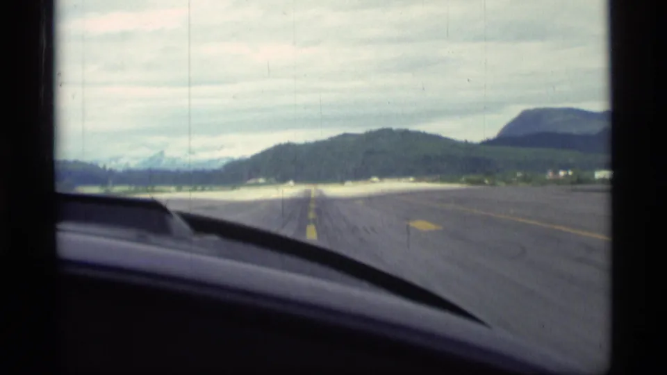 1977 Alaska home-movie frame showing a paved road from inside a moving vehicle, with green hills and mountains under cloud cover.