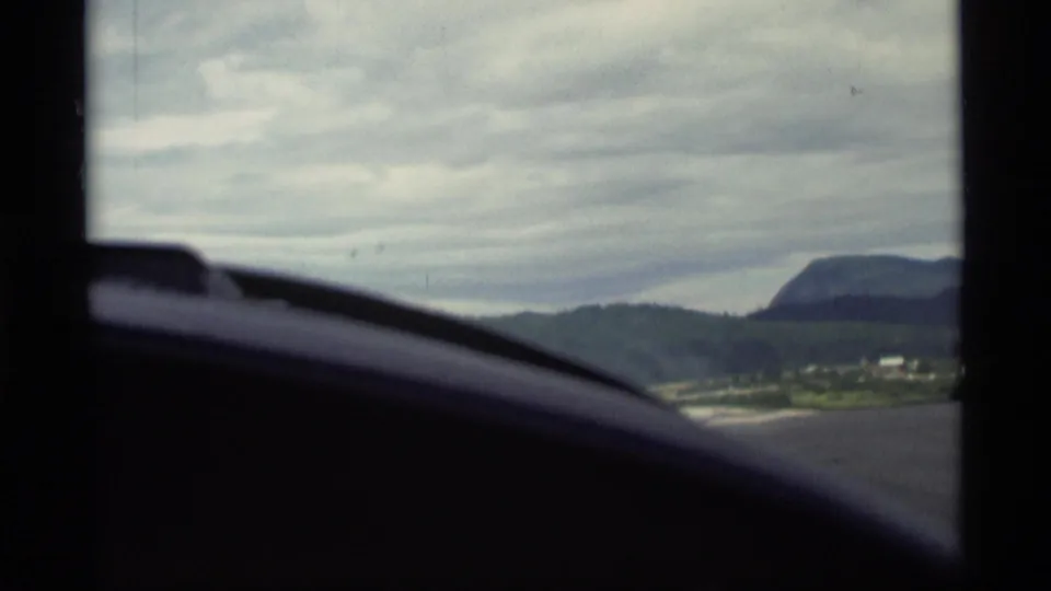 1977 Alaska travel frame looking past a dark vehicle interior toward roadside hills, cloudy sky, and a distant rounded mountain.
