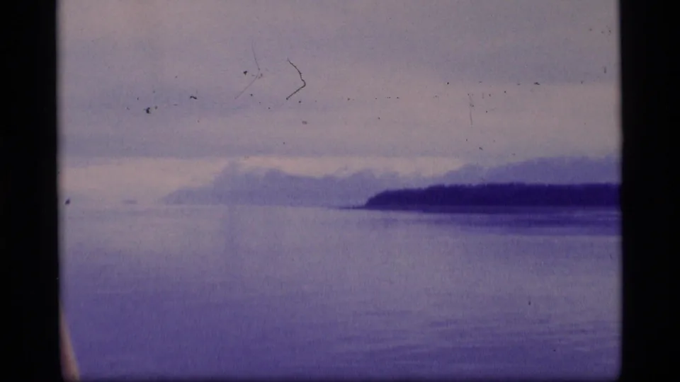 1977 Alaska coastal landscape frame with calm water, a forested shore, and hazy snow-touched mountains in the distance.