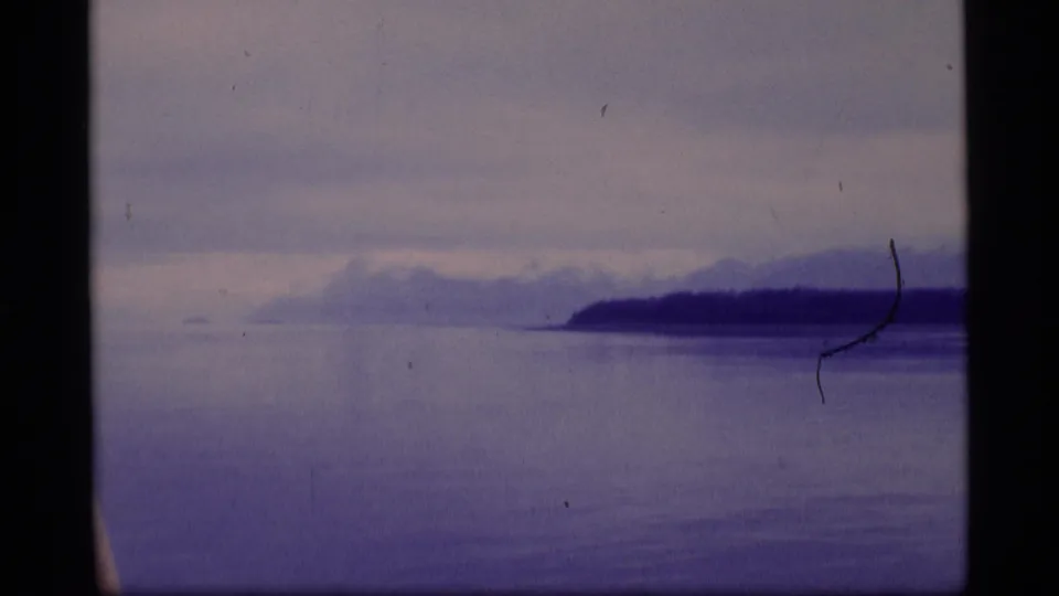 1977 Alaska home-movie frame of a bay or sound with dark shoreline, distant mountains, cloudy sky, and visible film wear.