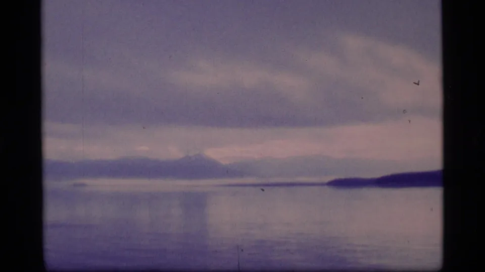 1977 8mm Alaska home-movie frame showing calm blue water, distant mountains, a low shoreline, and layered gray-purple clouds.