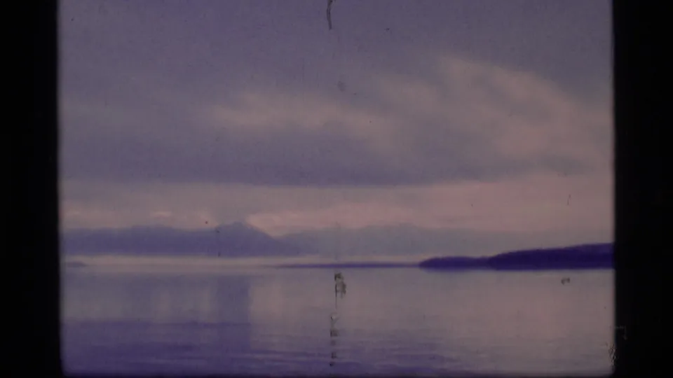 1977 8mm Alaska frame with broad water, faint mountain silhouettes, a dark shoreline at right, and soft cloud cover.