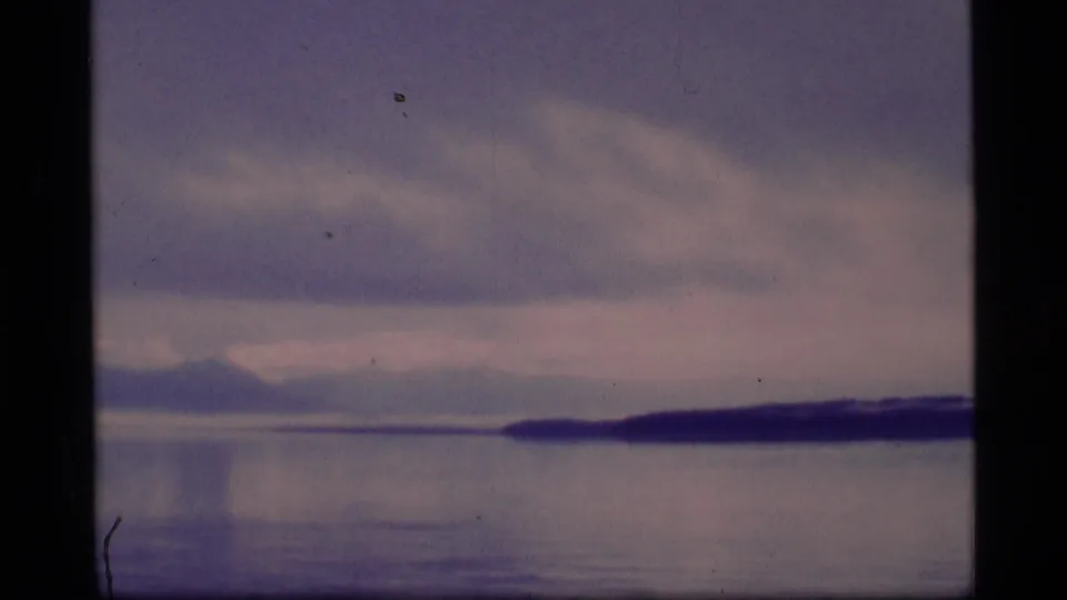 1977 8mm Alaska home-movie frame showing still water, low forested shoreline, distant mountains, and soft overcast light.