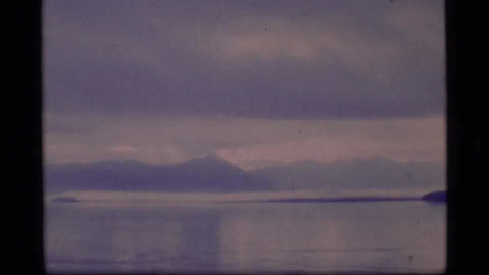 1977 8mm Alaska frame showing calm water, a distant mountain range, cloud layers, and a dark shoreline entering from the right.