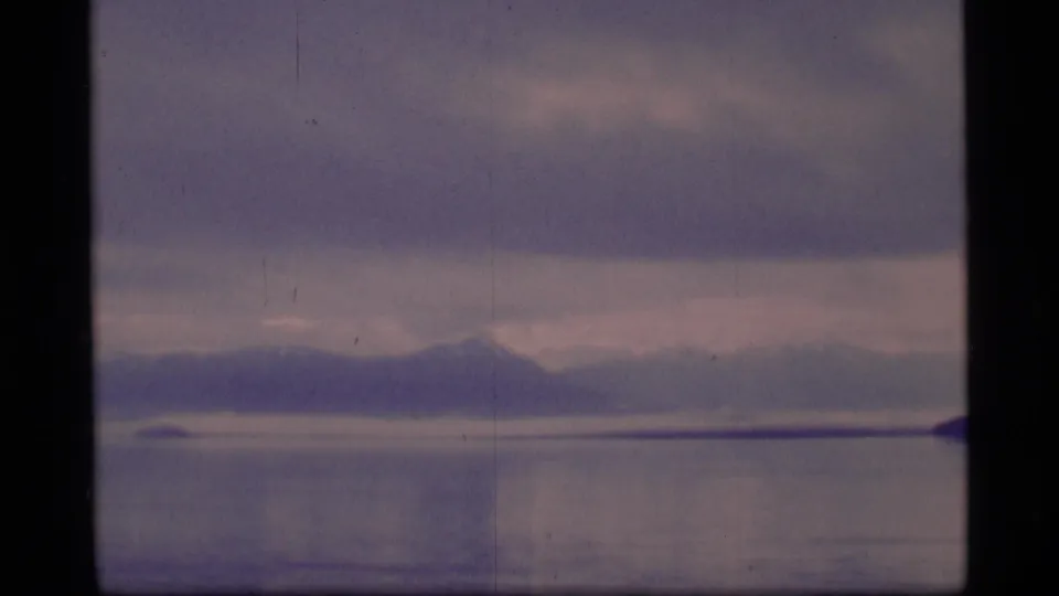 1977 Alaska 8mm frame showing a distant mountain ridge across calm water under an overcast sky.