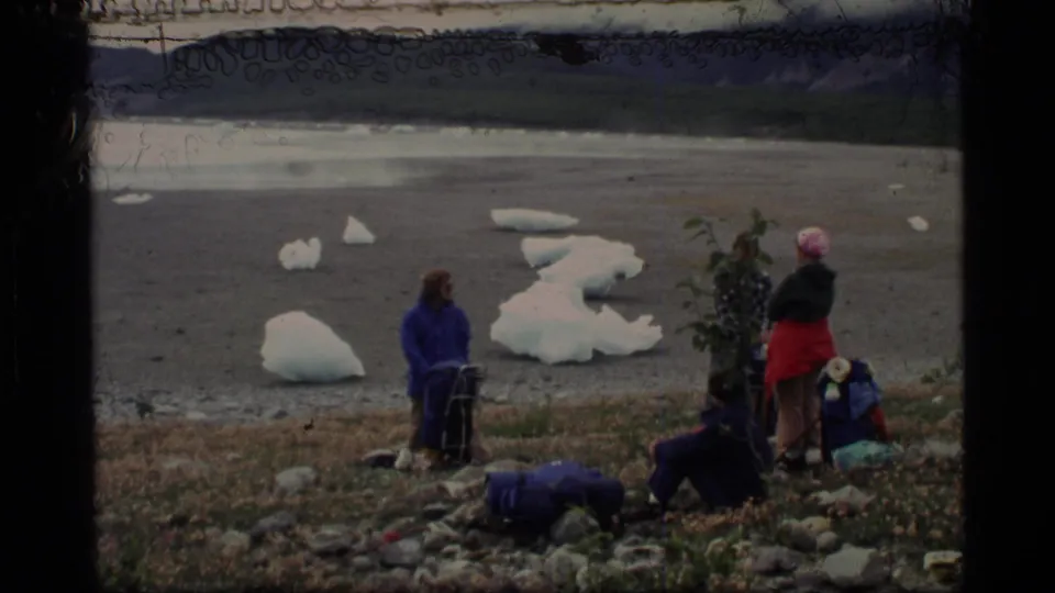 1977 Alaska lakeshore frame with adults in outdoor clothing near backpacks, floating ice, and distant mountains.
