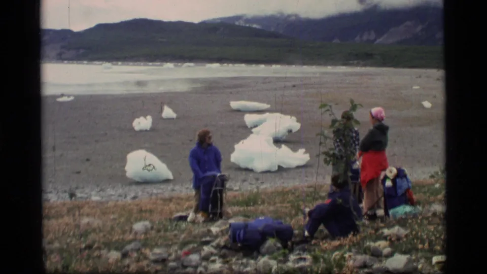 1977 Alaska home-movie frame of travelers resting with packs on a rocky glacial lakeshore.