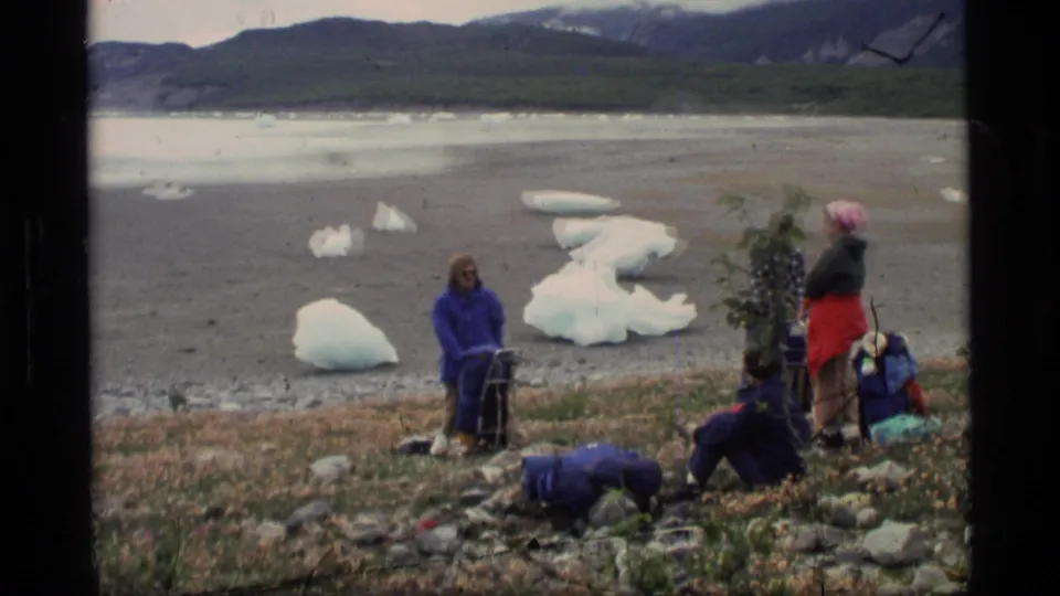 1977 Alaska 8mm frame showing adults resting beside gear on a glacial lake shore with mountains beyond.