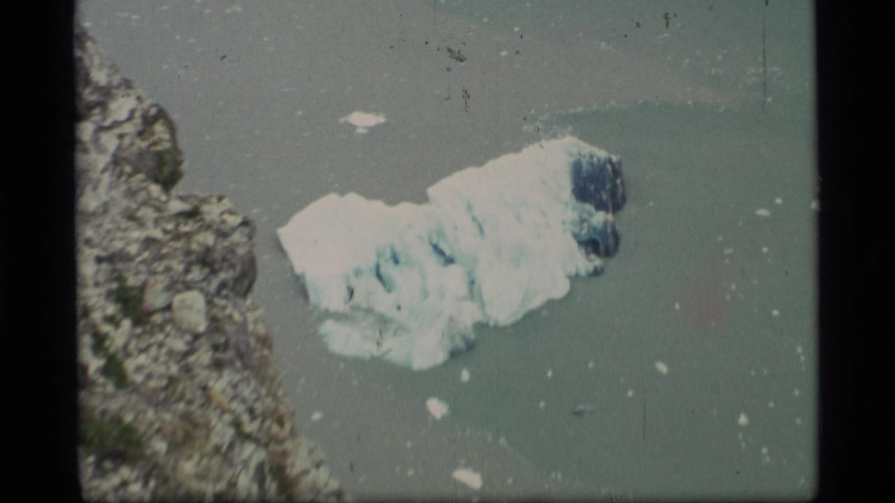 1977 Alaska 8mm frame showing a floating glacier chunk near a rugged coast, with small ice pieces in muted water.