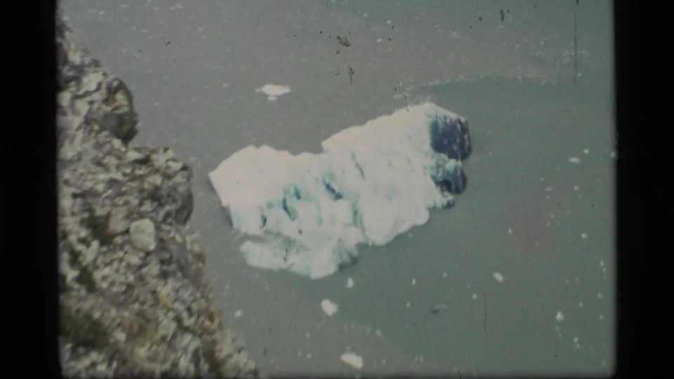 1977 Alaska 8mm frame showing a floating glacier chunk near a rugged coast, with small ice pieces in muted water.