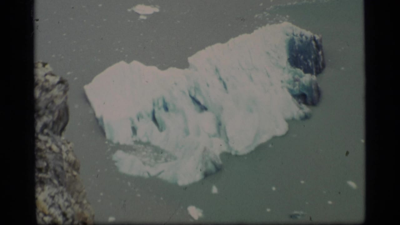 1977 Alaska home-movie frame with a closer view of a textured white and blue glacier chunk in icy coastal water.