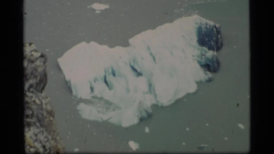 1977 Alaska home-movie frame with a closer view of a textured white and blue glacier chunk in icy coastal water.