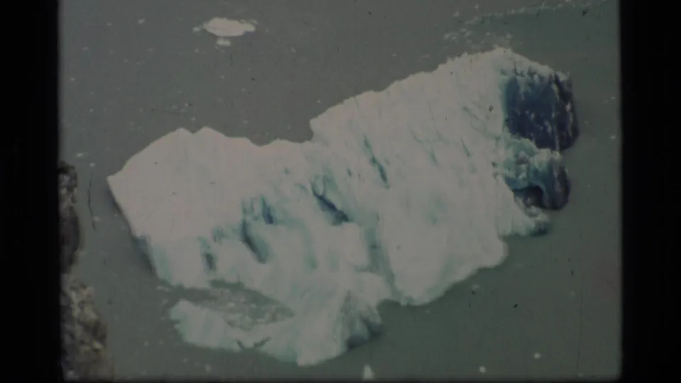 1977 Alaska home-movie frame of a glacier fragment drifting in coastal water, framed by 8mm borders and film wear.