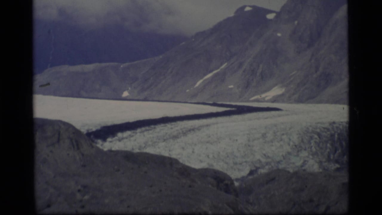 1977 Alaska 8mm frame showing a wide glacier below steep rocky mountains with snow patches and a cloudy sky.