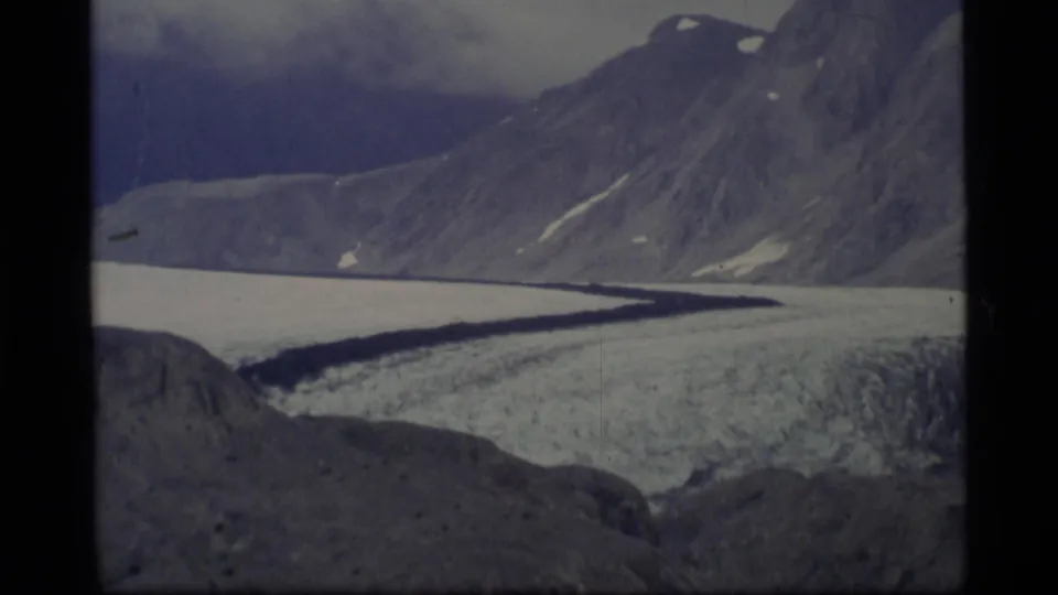 1977 Alaska 8mm frame showing a wide glacier below steep rocky mountains with snow patches and a cloudy sky.