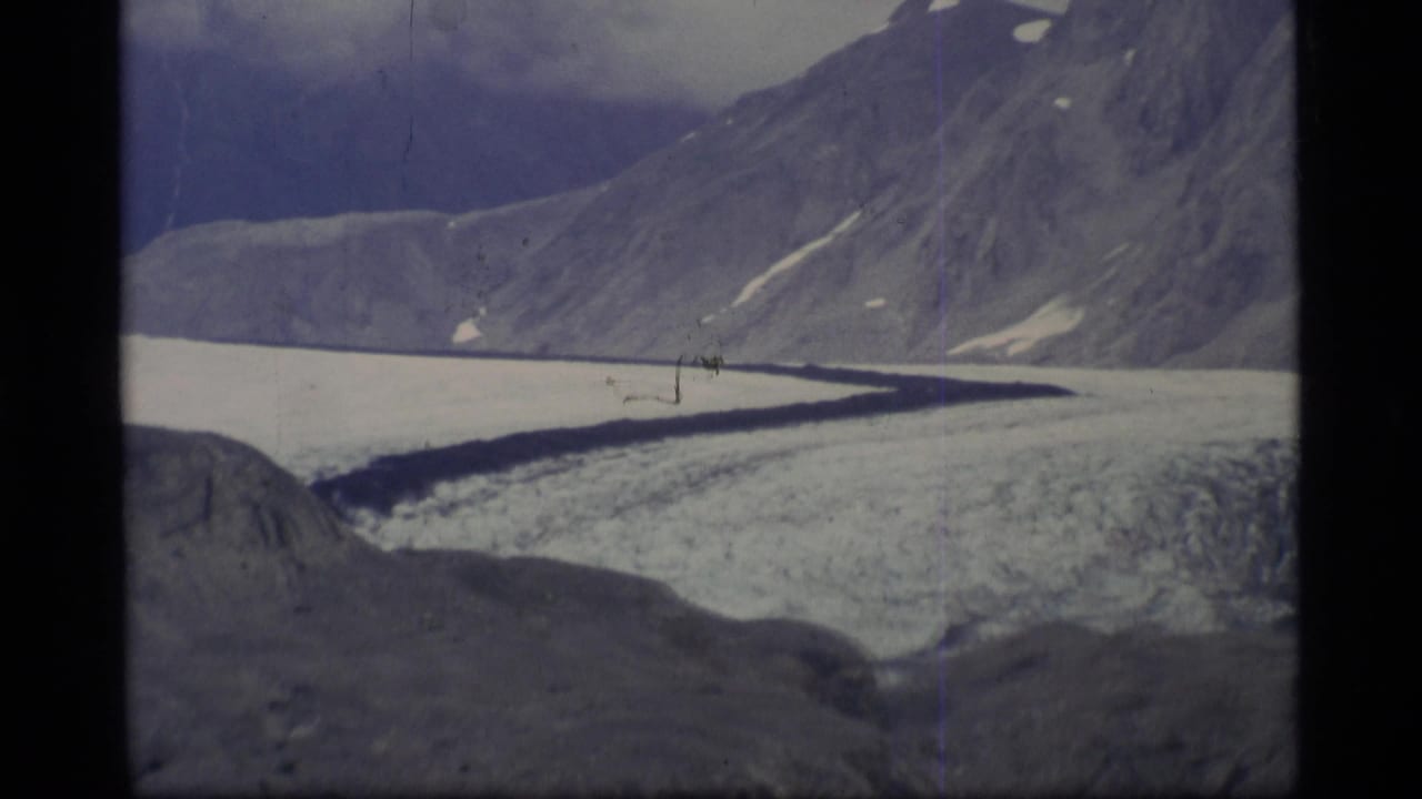 1977 Alaska home-movie frame of a glacier winding below steep mountain slopes and scattered snow patches.