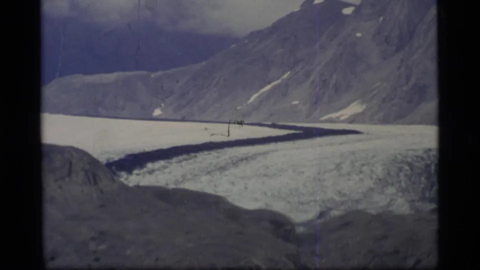 1977 Alaska home-movie frame of a glacier winding below steep mountain slopes and scattered snow patches.