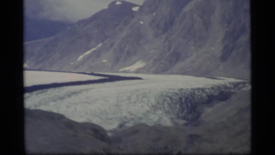 1977 Alaska home-movie frame showing the glacier edge below a rugged mountain slope in muted cloudy light.
