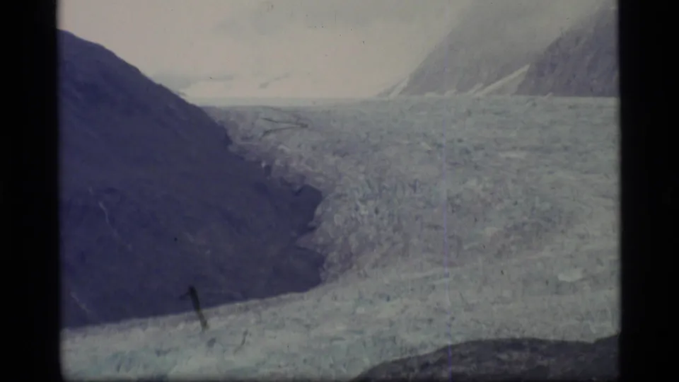 1977 Alaska 8mm frame showing a broad glacier running between dark mountain slopes under a pale sky.