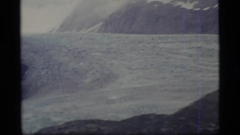 1977 Alaska 8mm frame showing a closer section of glacier ice, distant mountain slopes, and a dark foreground ridge.