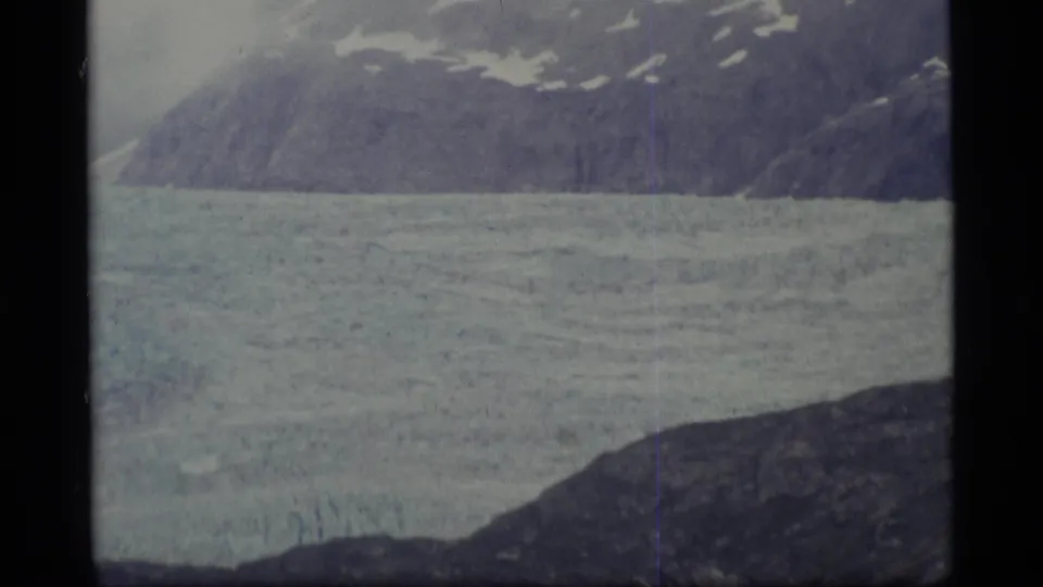 1977 Alaska 8mm frame of a glacier below a snow-dotted mountain slope, bordered by dark rock in the foreground.