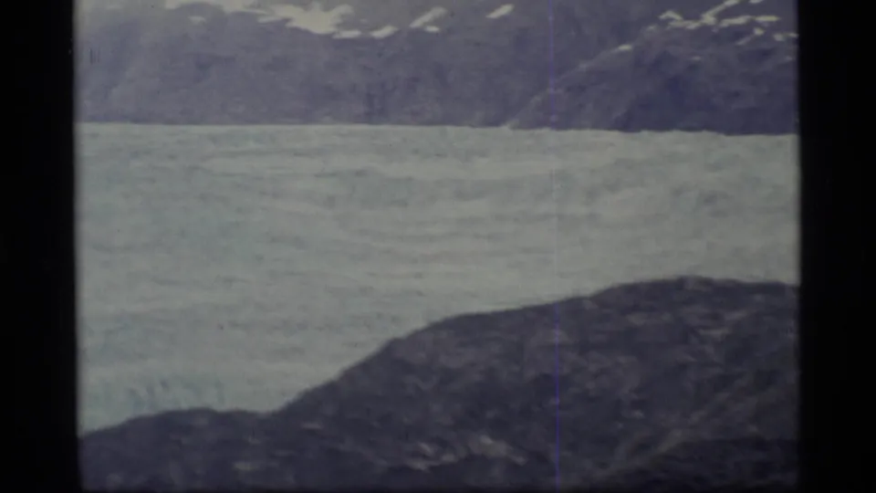 1977 8mm Alaska frame showing a pale glacier or frozen river beneath dark mountain slopes.
