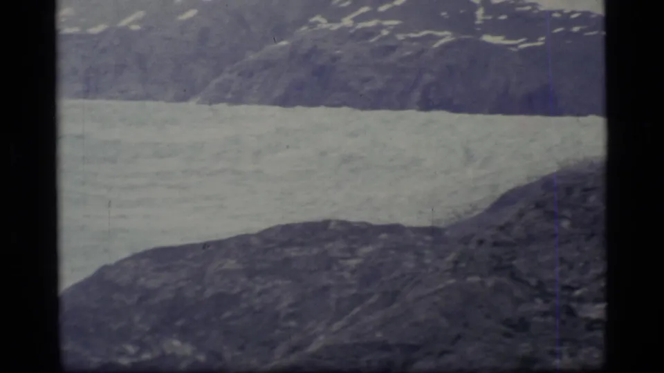 1977 Alaska home-movie frame with pale ice or river surface set against rugged dark slopes.