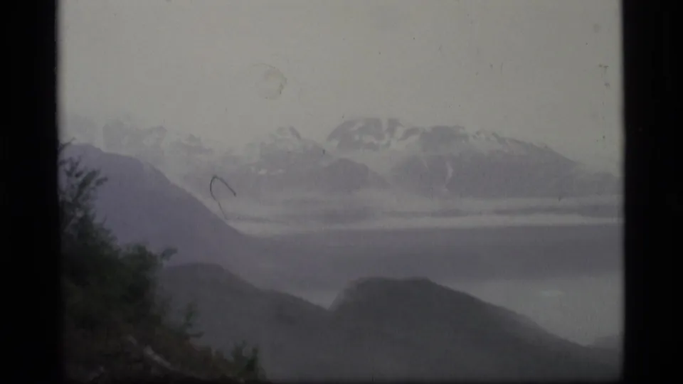 1977 8mm Alaska view of distant snow-capped mountains across a hazy body of water or valley.