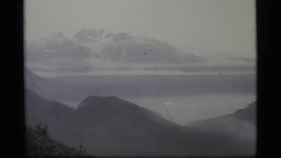 1977 Alaska frame showing a misty mountain range beyond dark hills and a pale waterline or valley floor.