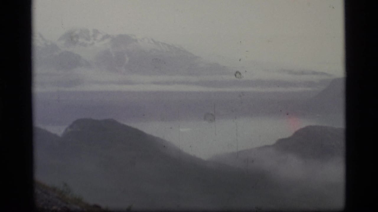 1977 Alaska 8mm frame showing distant snow-capped mountains, layered dark ridges, valley mist, and a pale hazy sky.