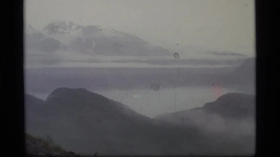 1977 Alaska 8mm frame showing distant snow-capped mountains, layered dark ridges, valley mist, and a pale hazy sky.