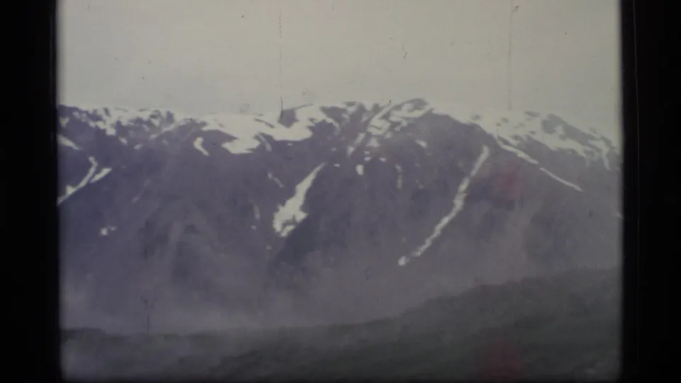 1977 Alaska 8mm frame of a snow-streaked mountain ridge under a pale sky, with haze drifting along lower slopes.